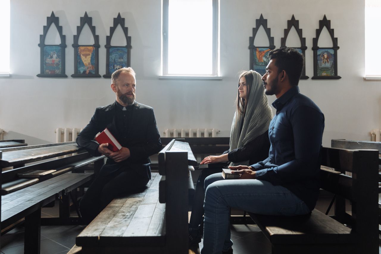 brand-01 A group of individuals discusses inside a church, seated on wooden benches.
